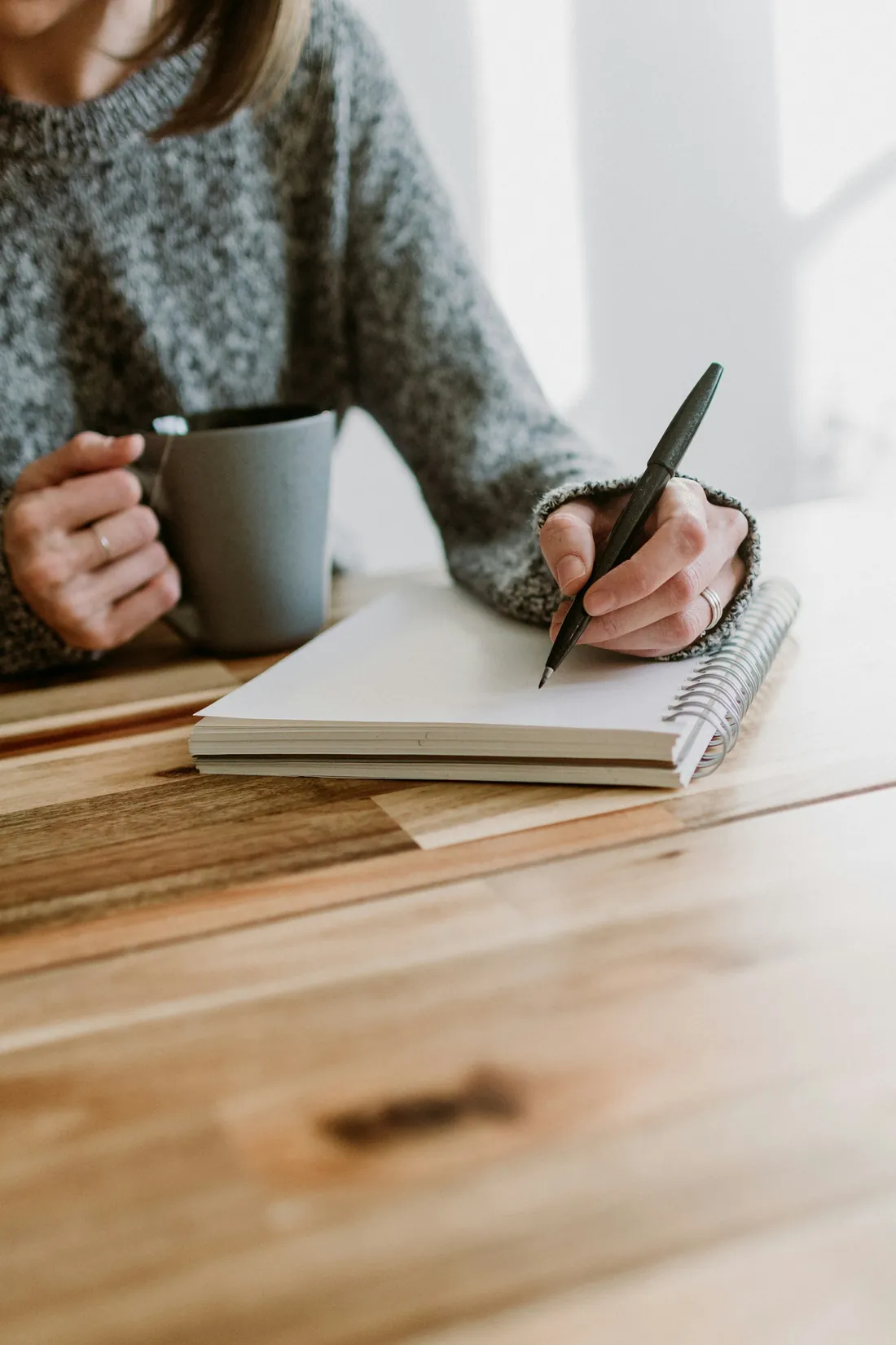 Une femme écrit dans un carnet, une tasse à la main, à une table en bois