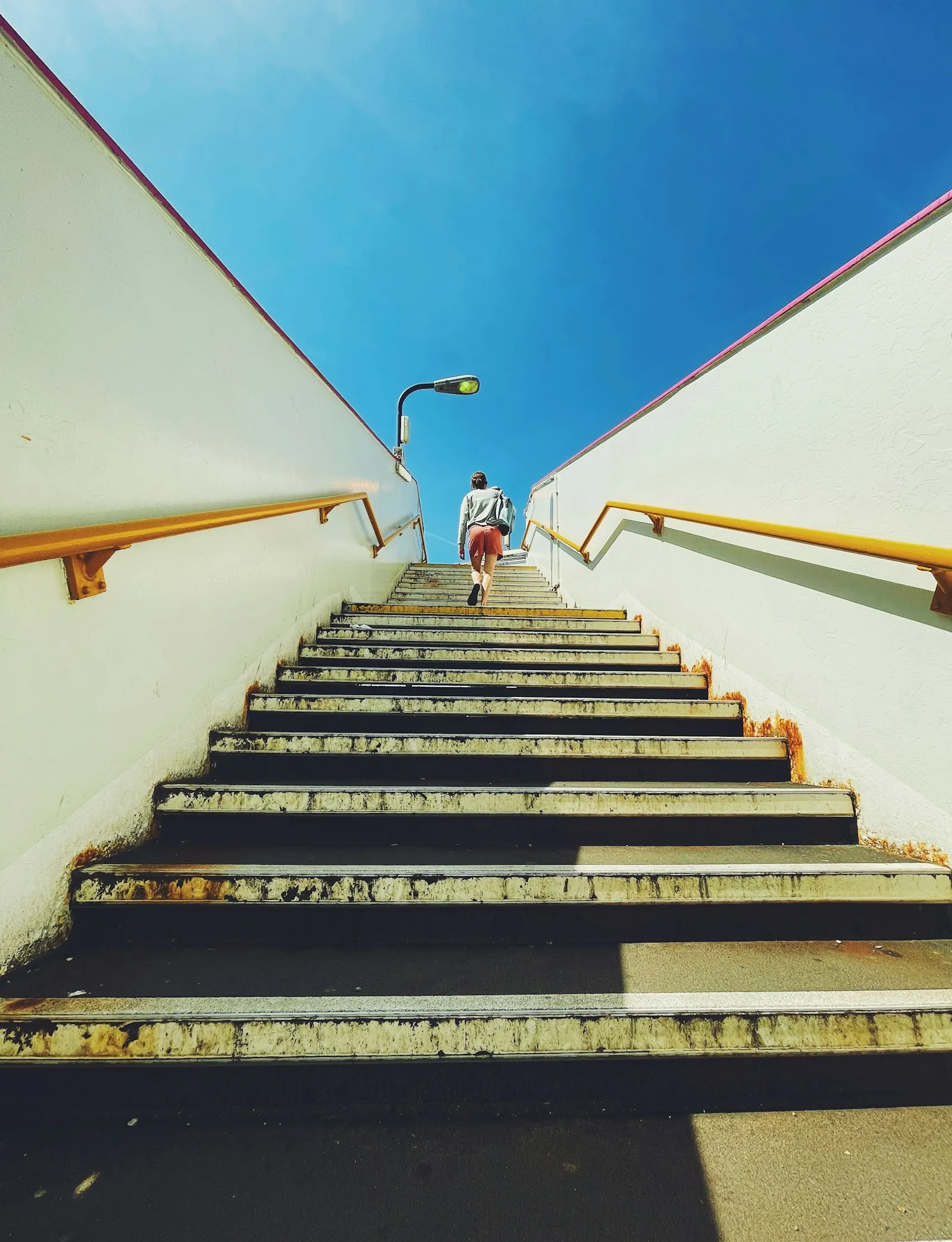 Une personne gravit un escalier aux rampes jaunes qui s’ouvre sur le ciel bleu